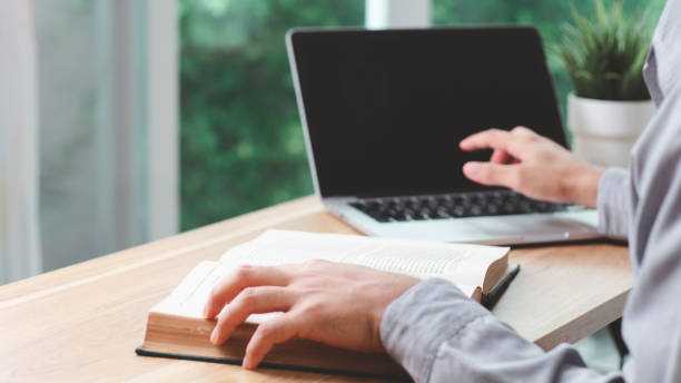 istock Close-up of a hand man reading a book, taking notes, and using a laptop to research information online. 2162194786
