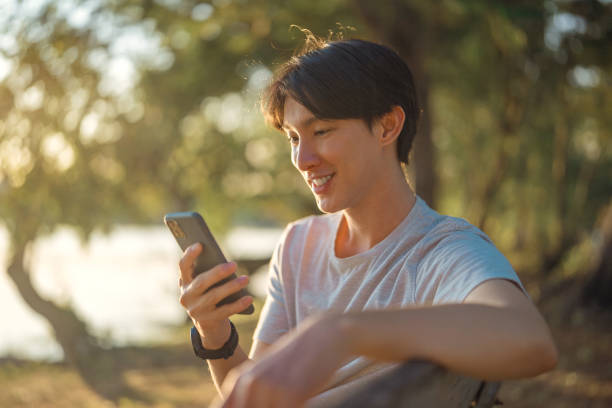 istock Happy young asian man using smartphone while sitting on bench in public park. 2162209301