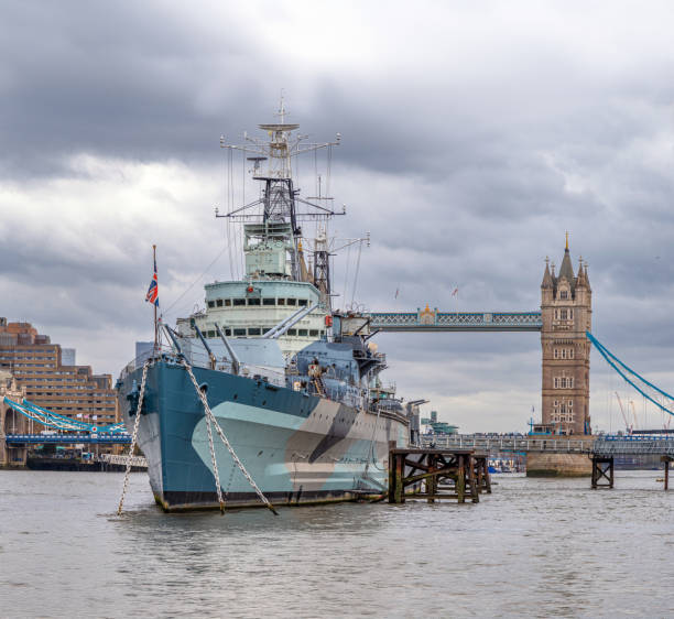 Front view of the floating war museum ship HMS Belfast docked on the River Thames with tourists visiting it and the tower bridge behind under a rainy cloudy sky.