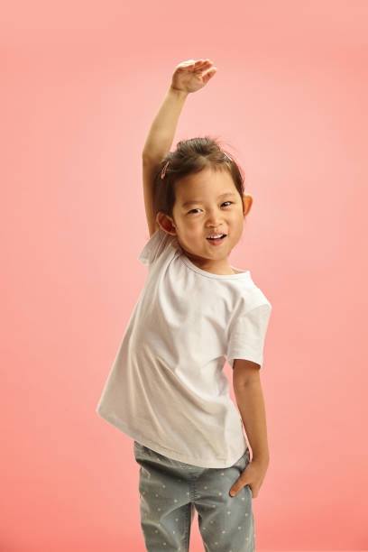 istock Little cheerful kid measures her height against and playful pulls her arm as high as possible symbolizing want to grow up standing on pink isolated. Concept of a girl's strength. 2162225078