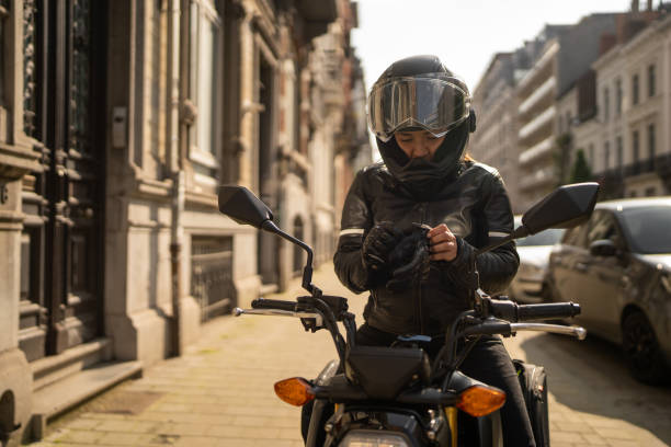 A young woman in a leather jacket is getting ready for a ride on her motorcycle. Surrounded by lush greenery, she smiles while putting on her gloves, embodying the spirit of adventure.