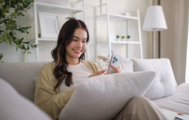 istock Cheerful young woman using mobile phone while sitting on couch at cozy home. 2162242125