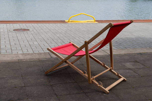 A red folding chair sits on a brick walkway near a body of water. A yellow metal barrier stands on the other side of the walkway.