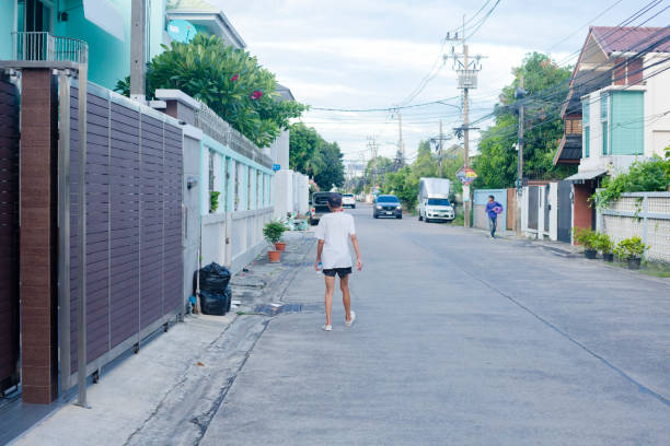 Thai senior man in shorts and tshirt is walking alone and slowly  in street of a local residential district in Bangkok Ladprao. Scene is in street Chockchai 4 Soi 56. Street is quiet traffic less frequented local community street with single homes and houses. Some houses have trees in front. Man is  a skinny person. He is passing by tall fences and gates of residential homes and buildings