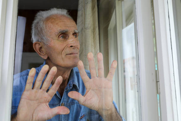 istock An elderly man wearing a blue denim shirt, standing by the window with both hands resting on it, gazing outside from his room. This image conveys a sense of introspection and solitude.