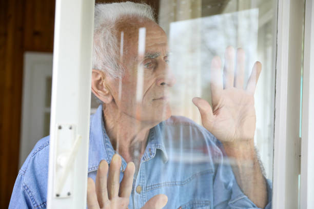 istock An elderly man in a blue denim shirt with both hands spread on the window, looking out from his room 2162282011