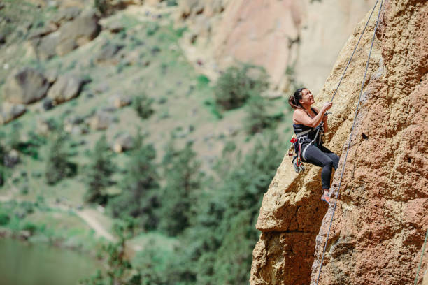 An active woman wearing protective gear uses a rope to help her climb up the side of Smith Rock in Central Oregon on a beautiful and sunny Spring day.