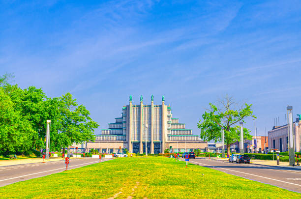 The drone footage captures the Monument to the Dynasty in Brussels, Belgium, showcasing its intricate Neo-Gothic architecture with pointed arches, detailed carvings, and a soaring spire. The monument, built to commemorate the Belgian monarchy, is set within a verdant landscape of manicured lawns and tree-lined pathways, enhancing its regal presence. The aerial perspective provides a sweeping view of the monument's symmetrical design and the surrounding park, highlighting the contrast between the historic structure and the green expanses. As part of the Royal Domain, this landmark symbolizes Belgium's royal history and serves as a significant cultural and historical attraction.