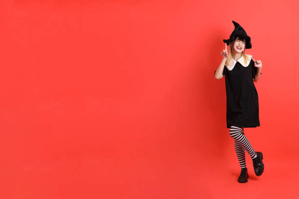 A young woman wearing a black witch's hat and intricate face paint in front of orange and black balloons, representing Halloween festivities and spooky themes.