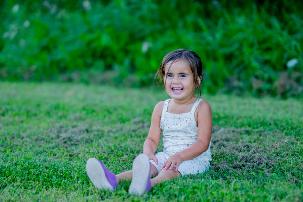 A sweet little girl sits outside in the grass on a warm summers day as she poses for a portrait.  She is dressed casually and smiling.