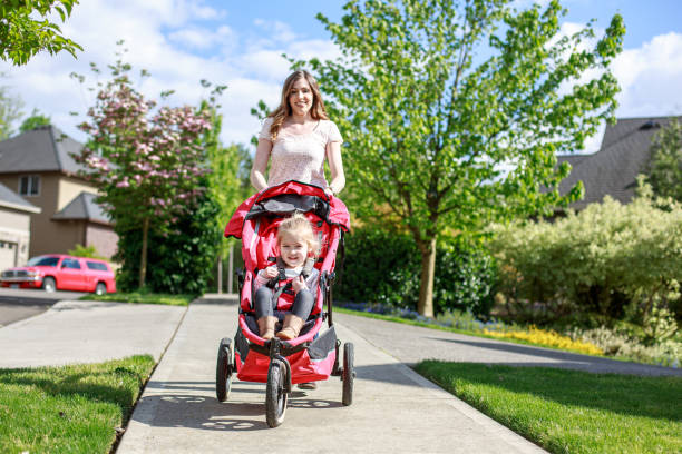 A Caucasian woman pushes her two year old daughter in a stroller on a neighborhood sidewalk, enjoying a beautiful Spring day in the sunshine.