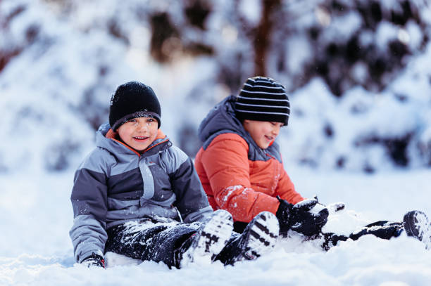 Two elementary age Eurasian brothers sit side by side in the snow and play.