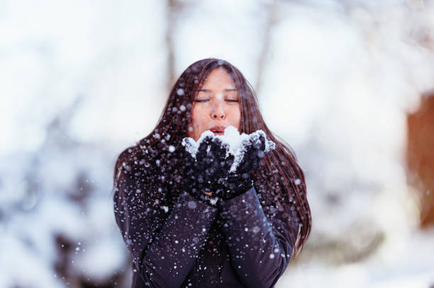 A beautiful woman of Pacific Islander descent blows snow flurries out of her hands while enjoying time outside on a Winter day.