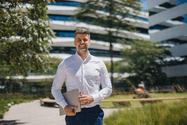 istock Smiling man holding laptop, modern architecture, trees 2162314904
