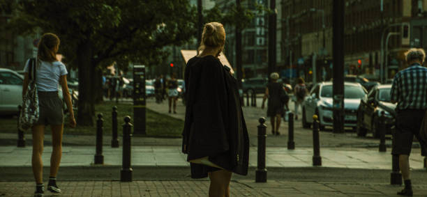 A back view of a blonde woman with tied hair, wearing a long dark coat, standing on a bustling street in Warsaw, Poland. The scene captures the vibrant city atmosphere just after sunset, with traffic lights glowing in the distance. Other pedestrians are also visible, adding to the lively urban setting.