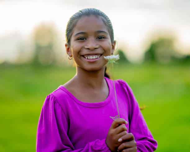 A young mixed race girl stands outside during sunset as she poses with a flower for a portrait.  She is wearing a comfortable pink shirt and smiling.