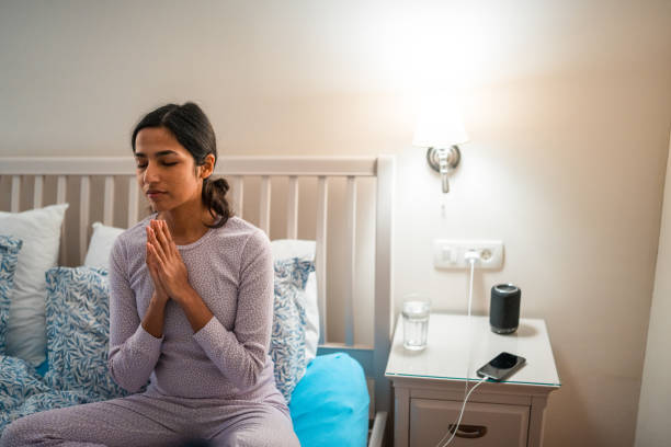 A serene Indian woman in comfortable nightwear practicing meditation on her bed with hands clasped, nighttime routine indoors by bedside with soft lighting.