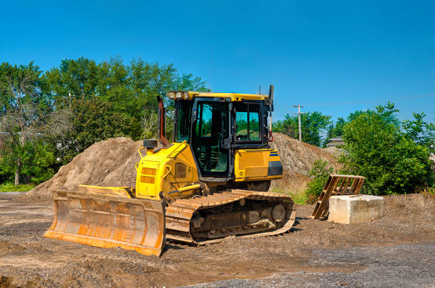 Yellow bulldozer with a front blade and caterpillar tracks. Its situated in an area with piles of dirt or sand, surrounded by trees and a clear blue sky.