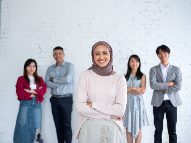 istock A diverse group of young professionals standing together against a white brick wall, with a confident young woman in a hijab smiling in the foreground 2162353224
