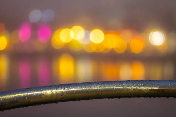 Metal railing with raindrops glistening, set against a vibrant, colorful city background at night. Urban lights create a dynamic, atmospheric scene highlighting the rain-soaked textures.