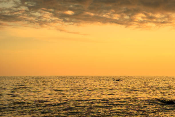 The sky is orange and the water is calm. The sun is setting and the clouds are in the background