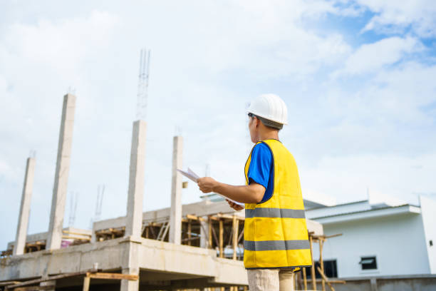 An architect, wearing a hardhat and safety vest, checks a laptop and house plan paper on a clipboard. Blueprints and construction documents lie nearby, emphasizing safety and precision in building. un arquitecto, con un casco y un chaleco de seguridad, revisa una computadora portátil y el papel del plano de la casa en un portapapeles. los planos y los documentos de construcción se encuentran cerca, enfatizando la seguridad y la precisión en la con