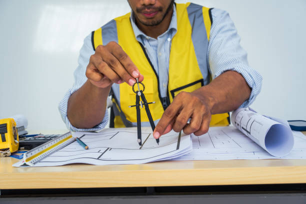 istock Asian architect works at a desk with house blueprints, checking plans on a laptop and clipboard, wearing a hardhat and safety vest, emphasizing precision and safety in office settings. 2162397285