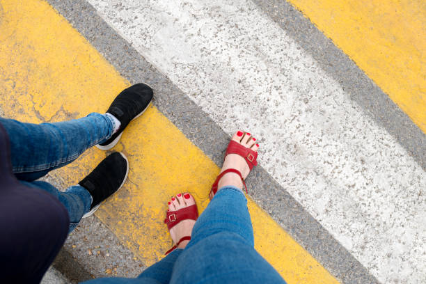 Feet in red sandals with red toenails and black shoes on a yellow-striped crosswalk.