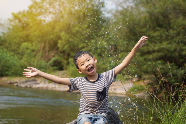 Asian boy taking a break from playing in a stream Sit on the rock, smiling brightly, arms outstretched to enjoy the splashing water happily. soft and selective focus.