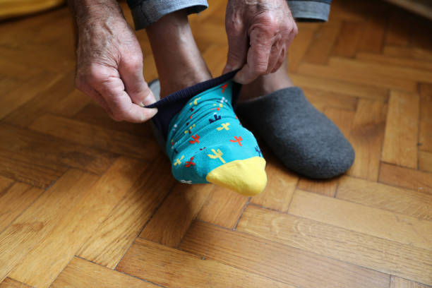 Close-up image of an elderly person putting on a colorful sock with cactus patterns. The person is seated on a wooden floor, with one foot partially in a gray slipper. The focus is on the hands pulling up the sock, highlighting the texture and details of the skin, the vibrant sock design, and the cozy home environment