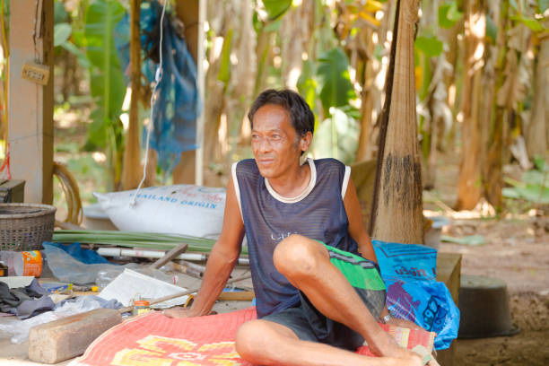 Mature thai farmer man is sitting on ground on  a plaid outside of rural farm and house in rural countryside of Phitsanulok province. In background are trees and banana trees. Scene in  ourter area of Amphoe Bangkratum with many banana plantations. Man is sitting together with some other banana plantation farm workers and farmer out of capture. Men are relaxing at late afternoon. Man has dark toned skin and is looking into distance. Next to him are several things as plastic waste, plastic bottle and a basket. Farmhouse is directly at banana plantation which is in background