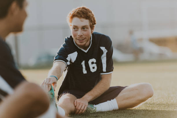 A young adult Caucasian man smiles to a teammate while sitting on a turf field, stretching his legs before playing in a soccer game.