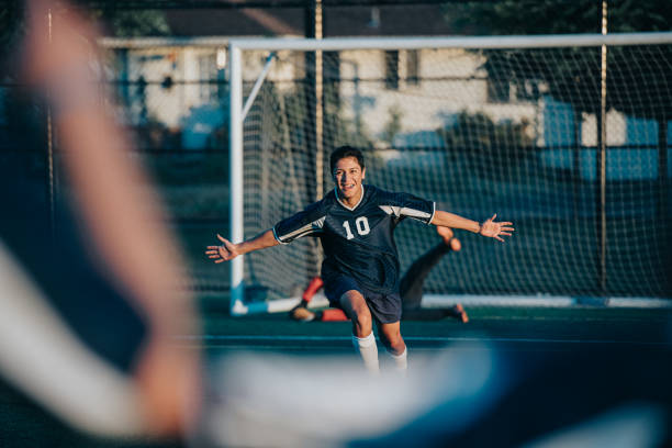 A young adult man of multiracial descent has his arms out in celebration after scoring a goal during an outdoor soccer game with fellow community members.