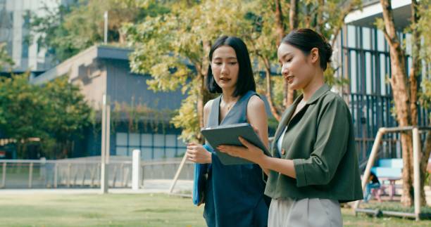 istock Two young Asian businesswomen discussing work while holds digital tablet and carries documents while walking outside office building. Sustainable business outdoor. 2162495280