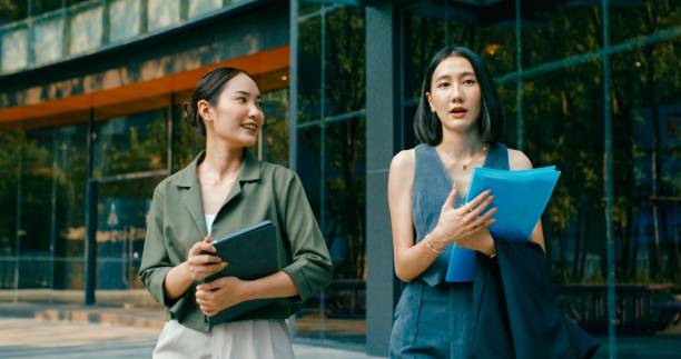 istock Two young Asian businesswomen discussing work while holds digital tablet and carries documents while walking outside office building. Sustainable business outdoor. 2162495457