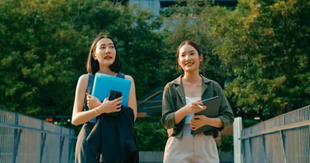 istock Two young Asian businesswomen discussing work while holds digital tablet and carries documents while walking outside office building. Sustainable business outdoor. 2162495578