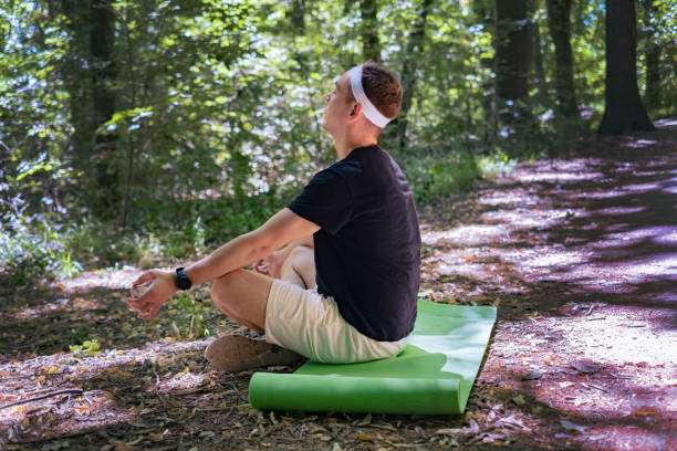 A bearded man sits in the forest on a mat, practicing meditation while listening to nature sounds.
