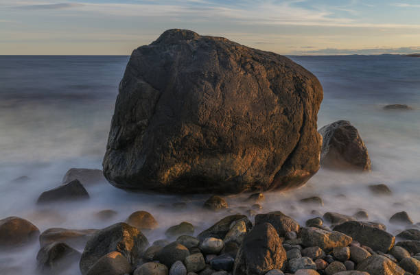 A large black rock rests on the seashore, silhouetted against a calm evening sky. The tranquil scene captures the serene beauty of dusk.