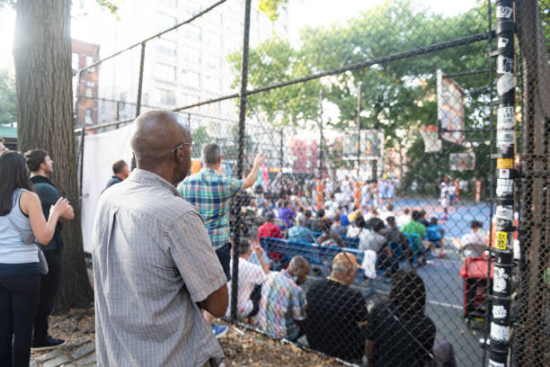 Seen from behind a small crowd of people watch a local basketball game from both sides of a chainlink fence in New York City.
