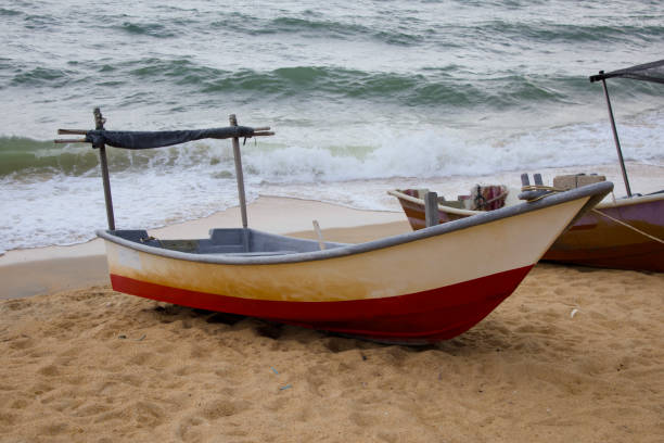 A vibrant red and white traditional fishing boat rests on the sandy beach of Pantai Teluk Lipat, Dungun, Terengganu. Equipped with wooden oars, it appears ready for a day at sea.