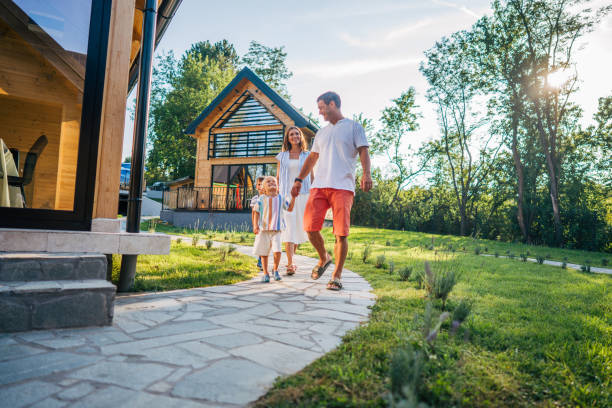 A family of four, consisting of a mom, dad, and their two sons, is enjoying a leisurely walk on a sunny day during their vacation. They are walking hand in hand along a stone pathway leading to their vacation home, which is surrounded by tall trees and lush greenery. The modern house in the background has large windows and a cozy, inviting appearance. The family looks happy and relaxed, with the children engaged and smiling. The warm sunlight and beautiful natural surroundings enhance the peaceful and joyful atmosphere of the scene.