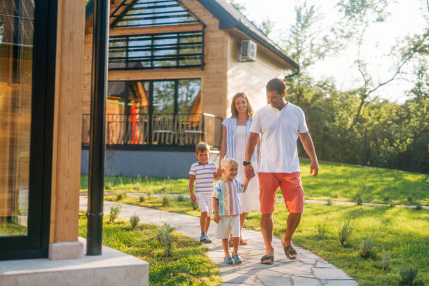 A family of four, consisting of a mom, dad, and their two sons, is enjoying a leisurely walk on a sunny day during their vacation. They are walking hand in hand along a stone pathway leading to their vacation home, which is surrounded by tall trees and lush greenery. The modern house in the background has large windows and a cozy, inviting appearance. The family looks happy and relaxed, with the children engaged and smiling. The warm sunlight and beautiful natural surroundings enhance the peaceful and joyful atmosphere of the scene.