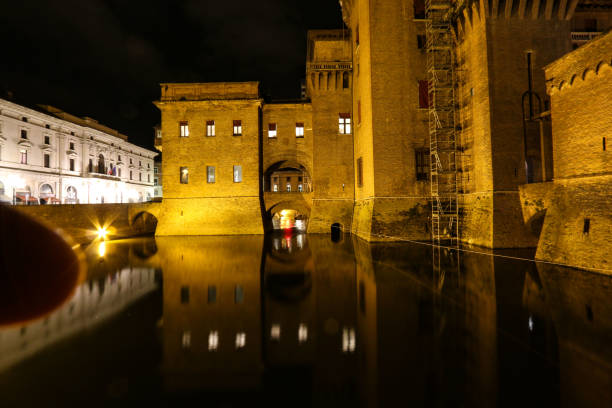 Ferrara, Emilia-Romagna, Italy - May 15th 2015 - Castello Estense and its Torre dei Leoni by night shows its massive walls emerging from the surrounding moat. Some windows are lit and reflect in the water