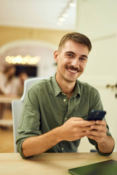istock A smiling man looking at the camera while using a mobile phone and sitting at the office. 2162560409