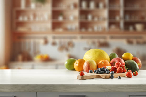 Blurred kitchen interior and fresh fruit on wooden cutting board on white countertop and space for food and drink and products and objects. Natural daily light coming through the window.