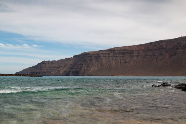 Dark brown to black color of the cliffs on the north of Lanzarote. Shallow water of a beach. Blue sky with white clouds. Caleta del Sebo, La Graciosa, Canary Islands, Spain.