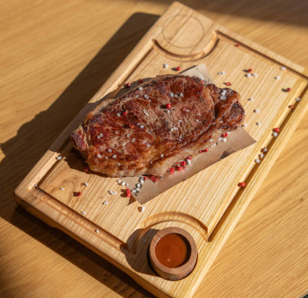 a perfectly cooked steak sits on a wooden board, seasoned with salt and peppercorns. There is also a small dish of sauce on the side