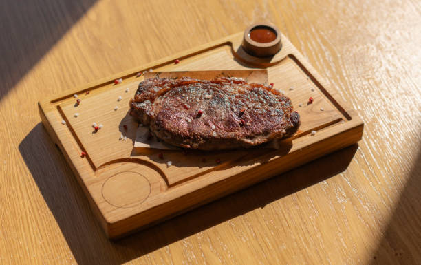 a perfectly cooked steak sits on a wooden board, seasoned with salt and peppercorns. There is also a small dish of sauce on the side