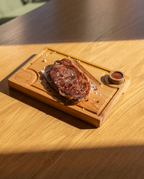 a perfectly cooked steak sits on a wooden board, seasoned with salt and peppercorns. There is also a small dish of sauce on the side