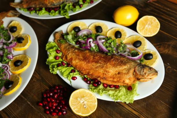 A fried fish dish with lemon slices, pomegranate seeds, and red onion on a white plate.